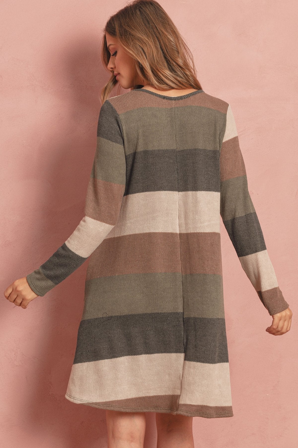 A woman poses in the Riah Fashion Long Sleeved Rib Stripe Pocket Dress in Charcoal/Mocha against a pink textured background.