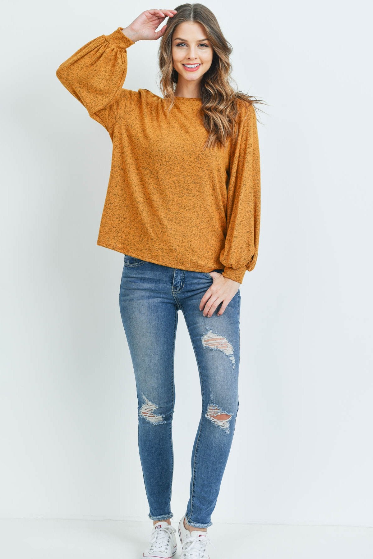 A woman models a Riah Fashion Puff Sleeved Boat Neck Two Toned Brushed Hacci Top with ripped jeans and brown boots on a concrete floor against a plain backdrop, accessorized with a long necklace.