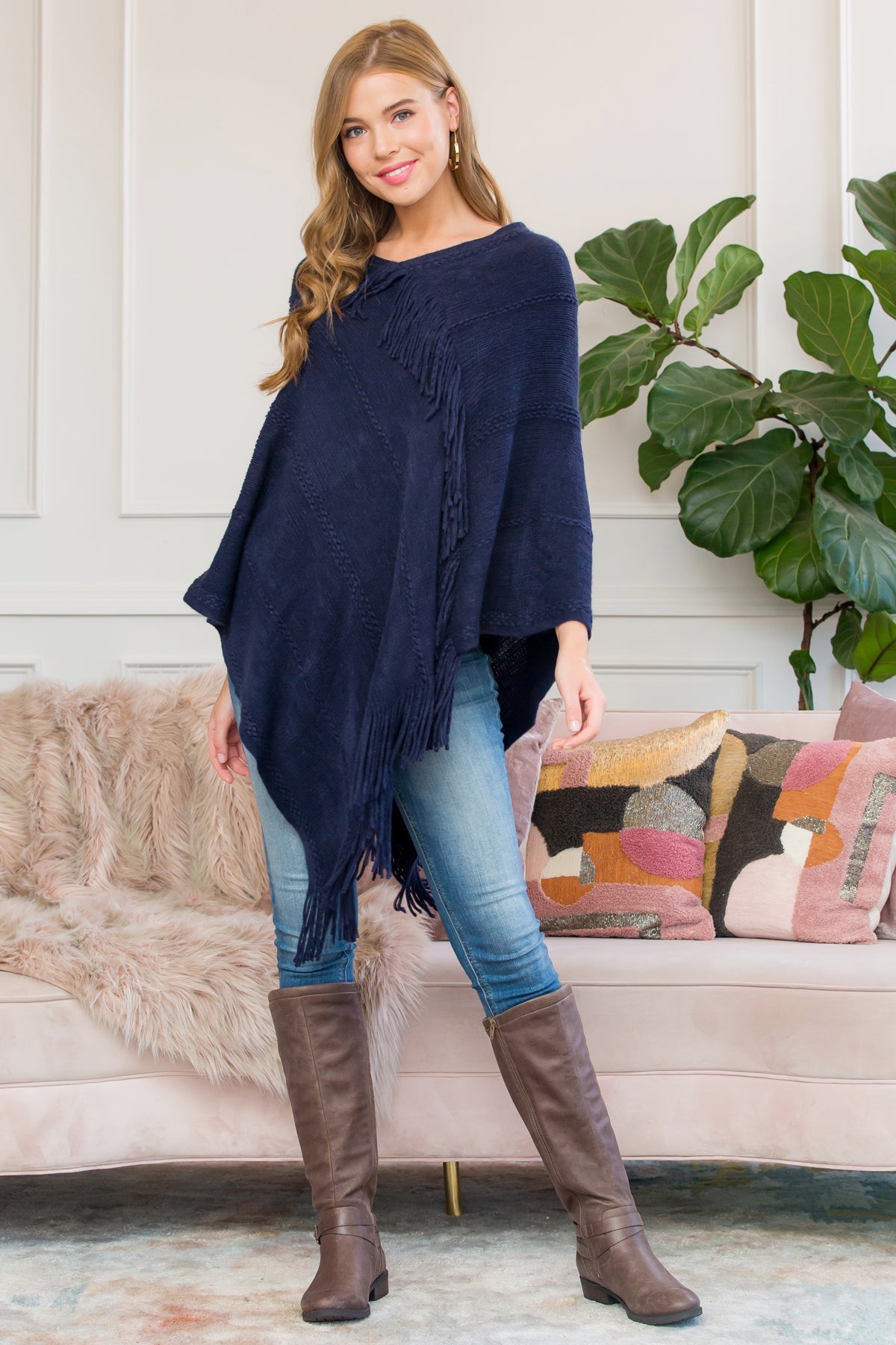 A person stands wearing a Riah Fashion Asymmetric Fringe Poncho in one color, paired with jeans and brown boots, smiling while holding the poncho. In the background are a plant and a sofa with cushions.