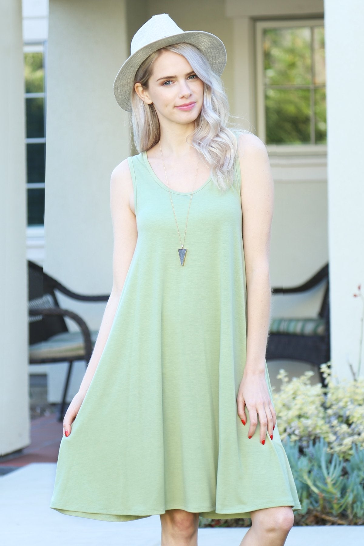 A person with long, light-colored hair wears a Sleeveless Flared Pocket Dress in light green by Riah Fashion outside, holding the dresss sides along with a triangular pendant necklace.