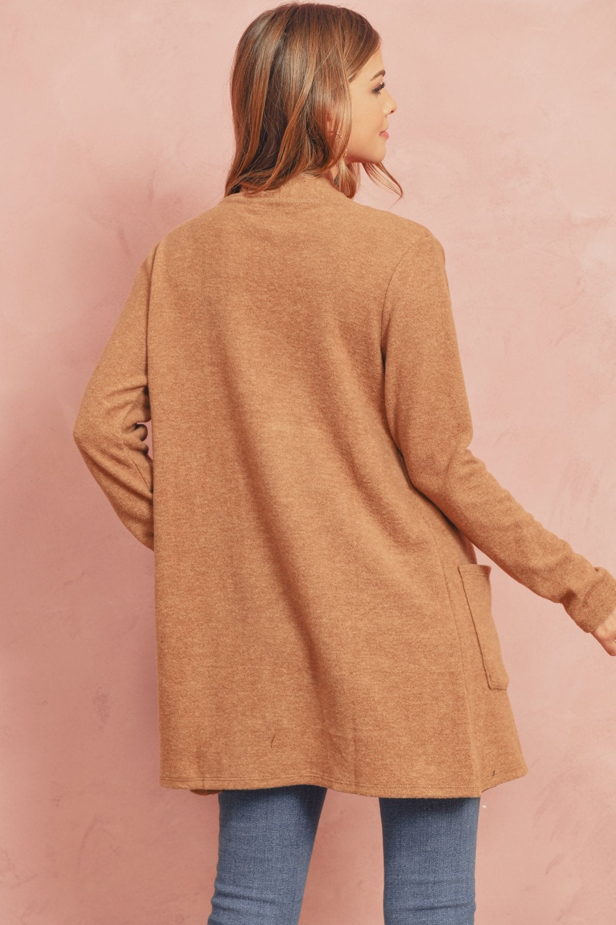 A woman in a Brushed Melange Open Front Cardigan by Riah Fashion and ripped jeans stands against a pink background, hands in her pockets.