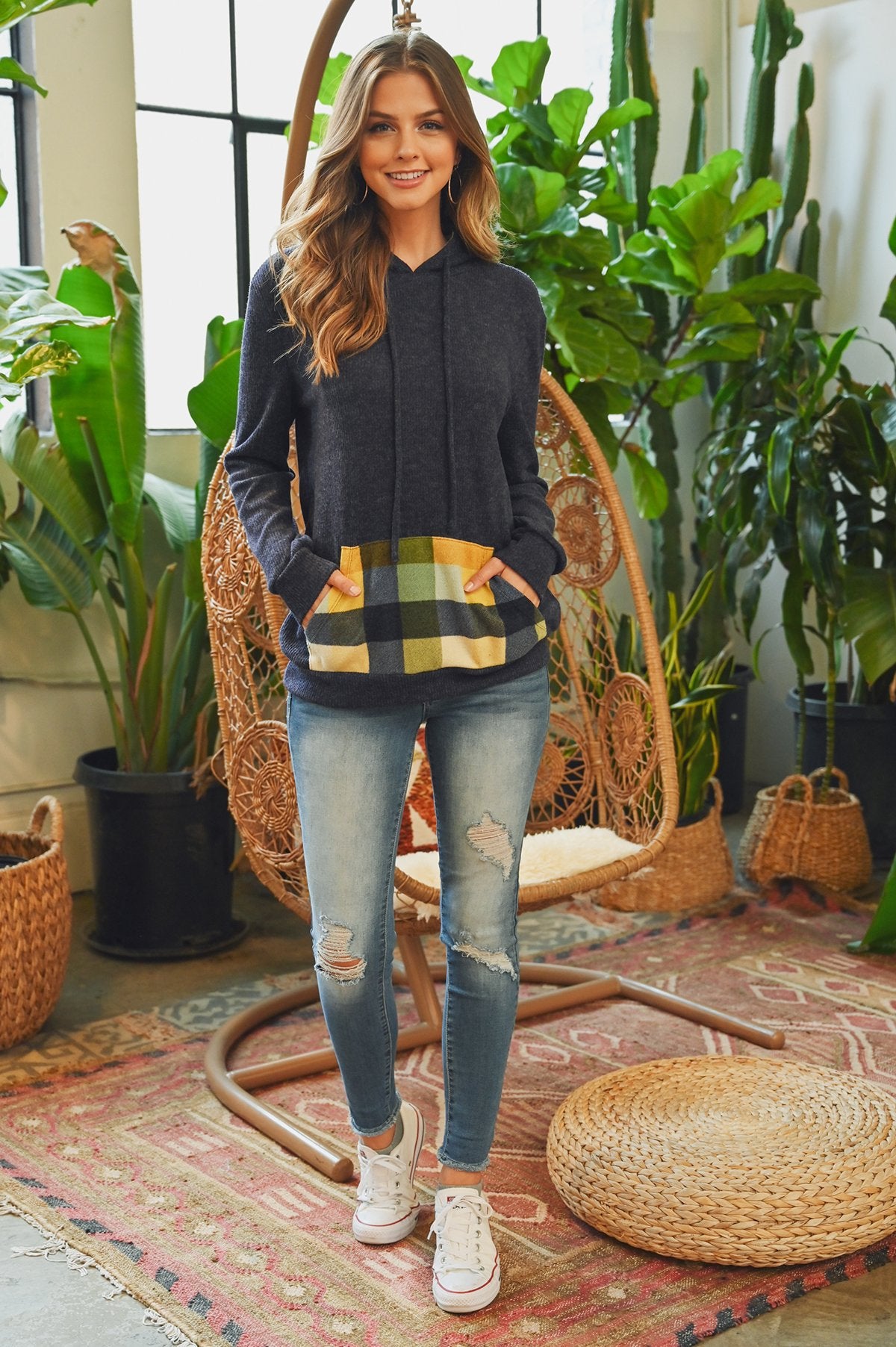 Someone wearing the Riah Fashion Hacci Brushed Plaid Kangaroo Pocket Hoodie stands indoors with plants in the background.