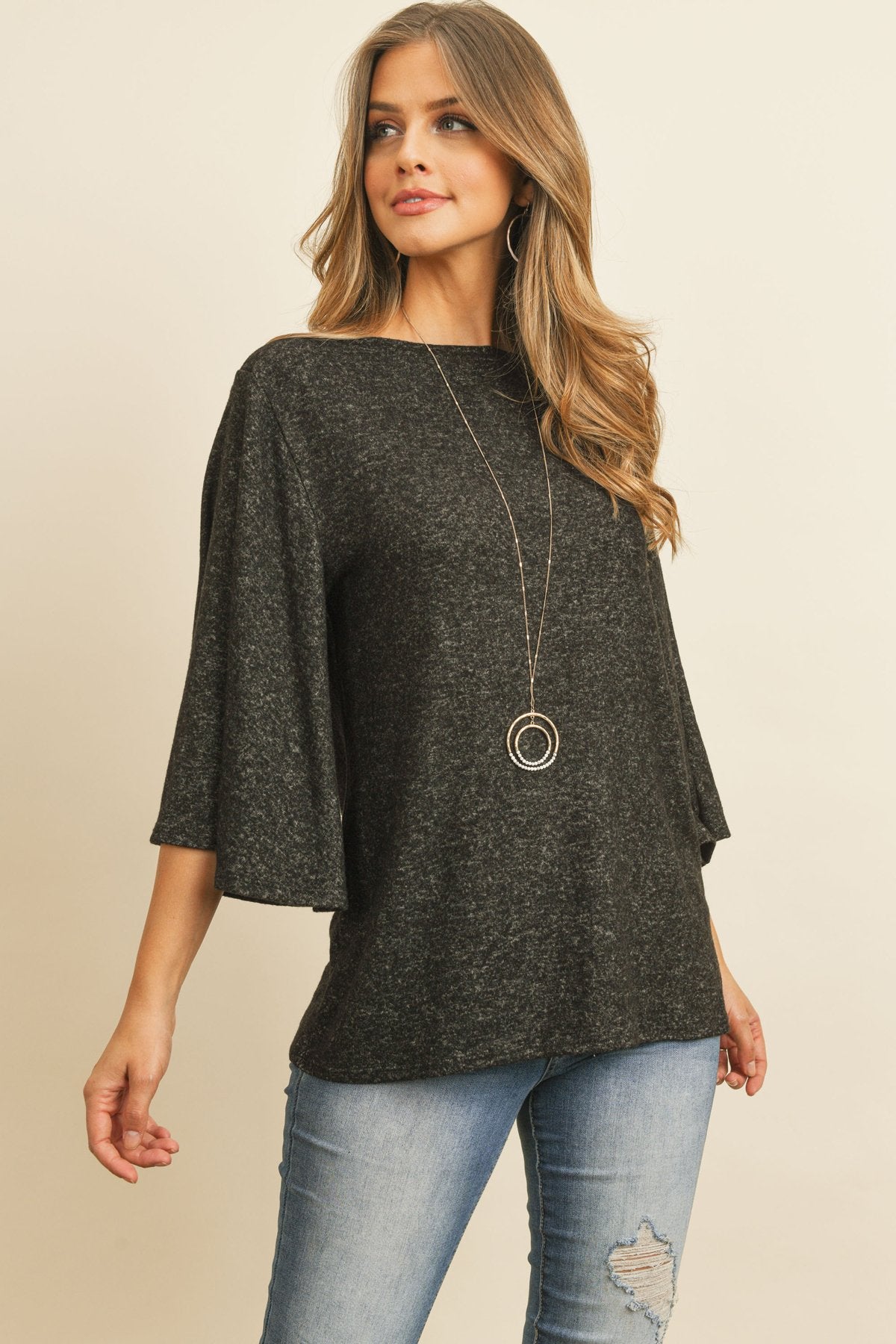 A person in a Riah Fashion Boat Neck Bell Sleeve Solid Hacci Brushed Top and ripped jeans stands against a neutral background, accessorized with hoop earrings and a long necklace.