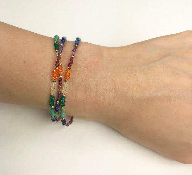 The Chakra Triple Strand Bracelet by Gena Myint is a multicolored beaded bracelet with three strands, featuring a gold clasp and chain extender.