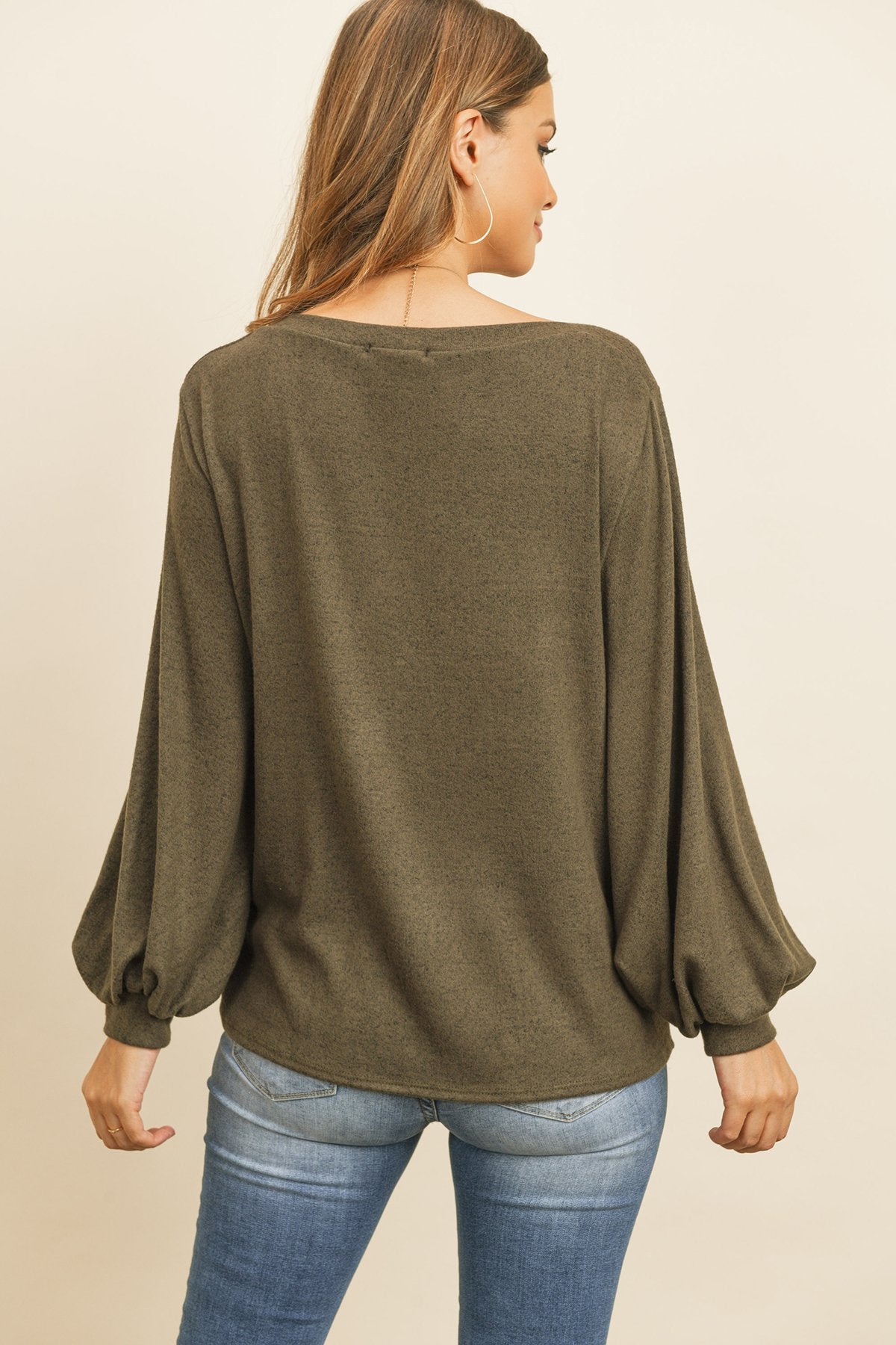 A woman models a Riah Fashion Puff Sleeved Boat Neck Two Toned Brushed Hacci Top with ripped jeans and brown boots on a concrete floor against a plain backdrop, accessorized with a long necklace.