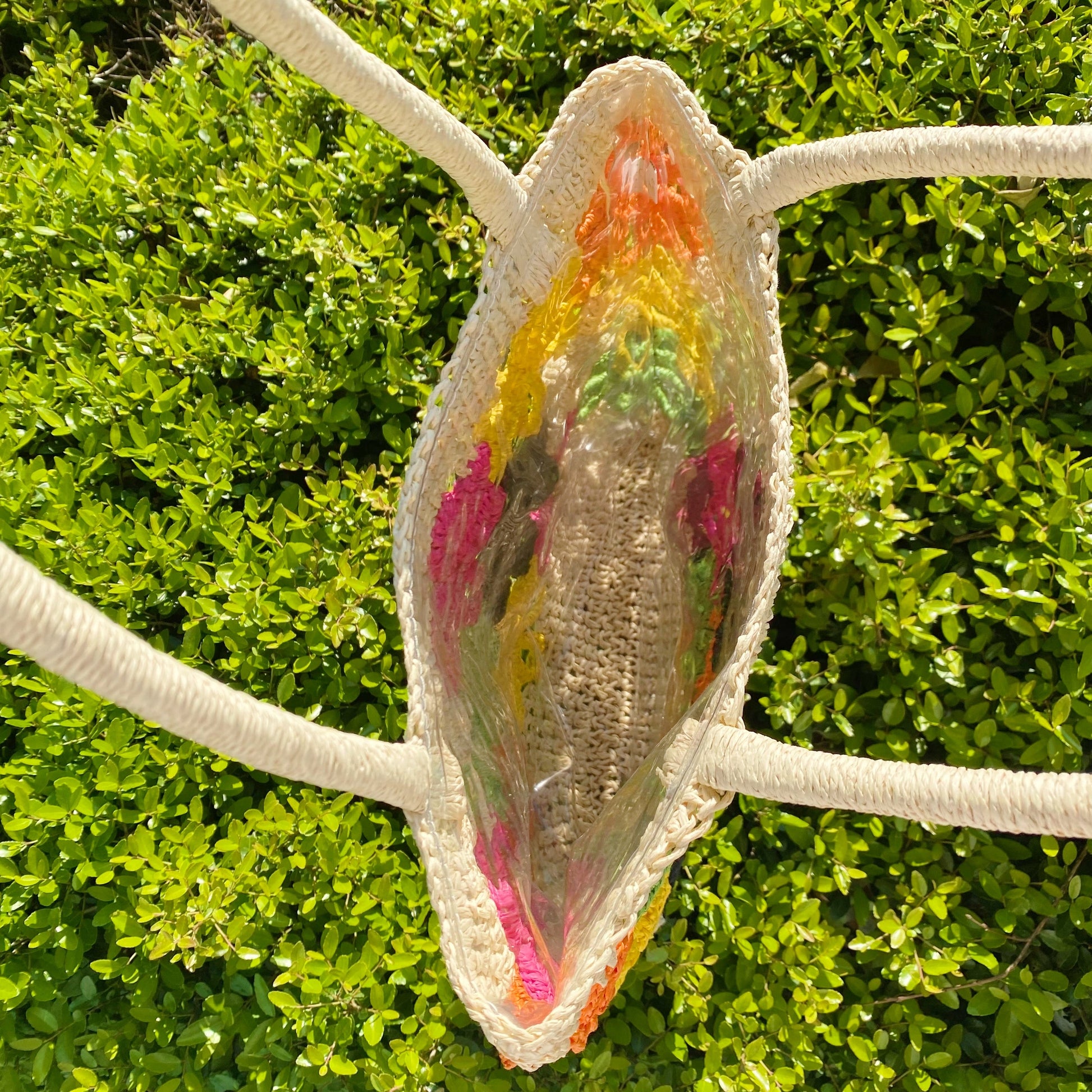 The Hand Made Flower Power Tote by Ellison + Young, featuring colorful floral crochet patterns in orange, green, pink, black, and yellow, rests on dense green foliage.