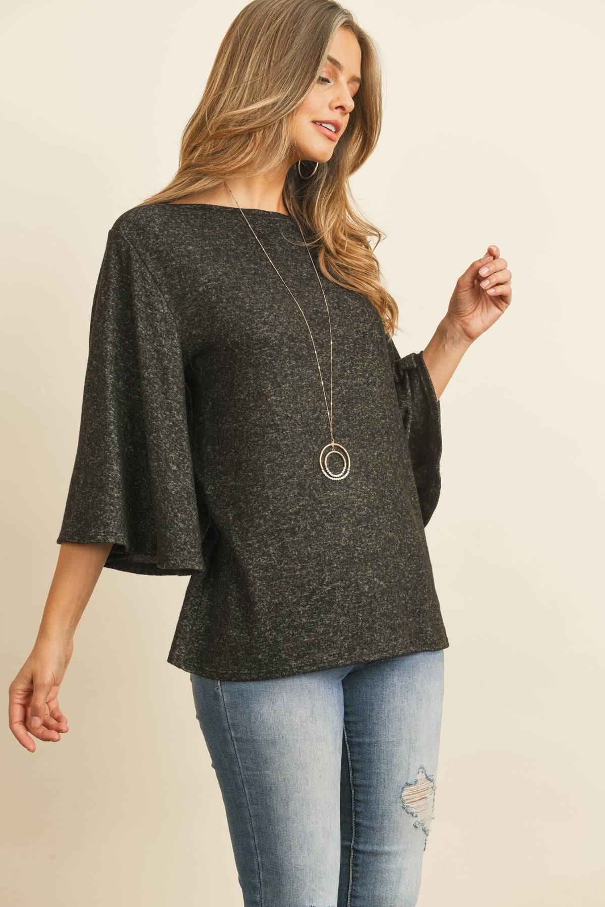 A person in a Riah Fashion Boat Neck Bell Sleeve Solid Hacci Brushed Top and ripped jeans stands against a neutral background, accessorized with hoop earrings and a long necklace.