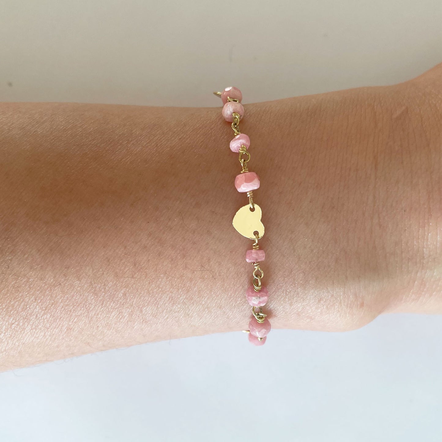 The Pink Rhodocrosite and Heart Charm Bracelet by Gena Myint is a gold chain bracelet with pink stones, three heart-shaped charms, and an adjustable clasp.