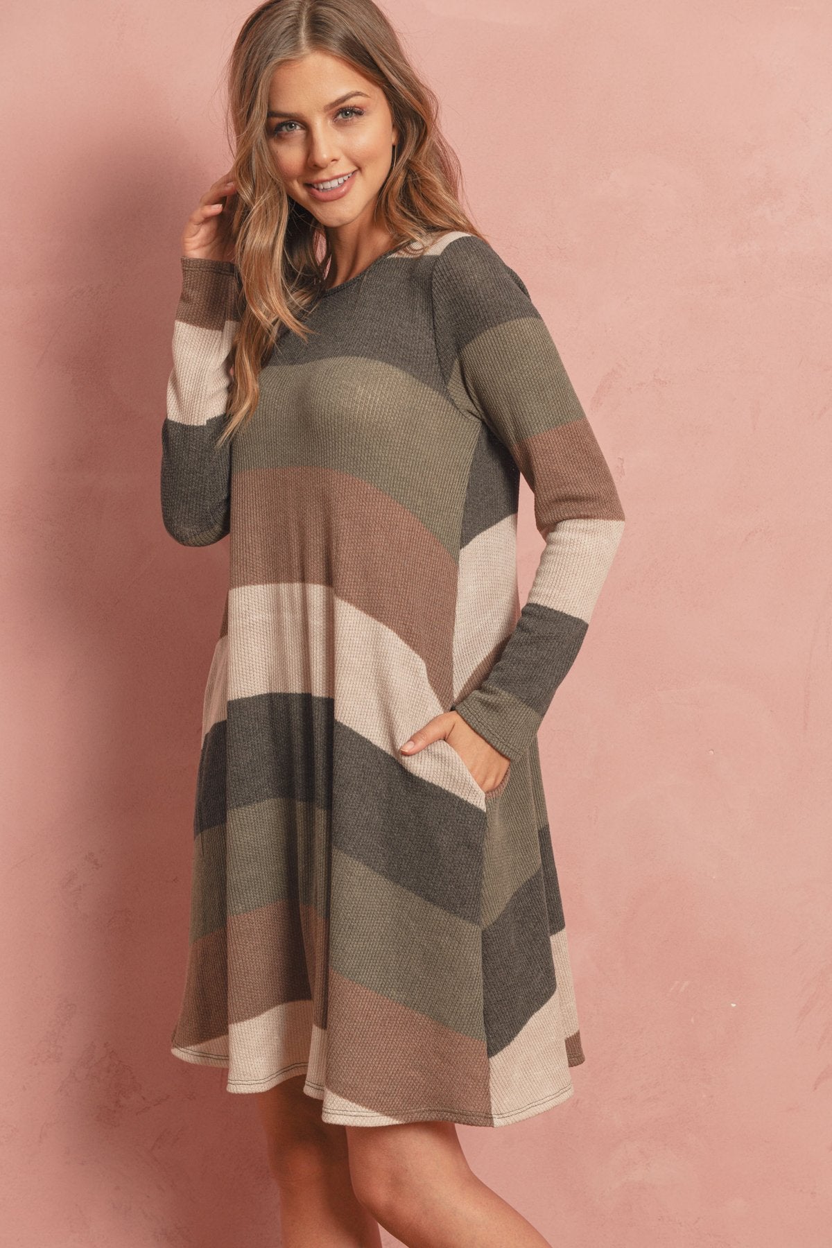 A woman poses in the Riah Fashion Long Sleeved Rib Stripe Pocket Dress in Charcoal/Mocha against a pink textured background.