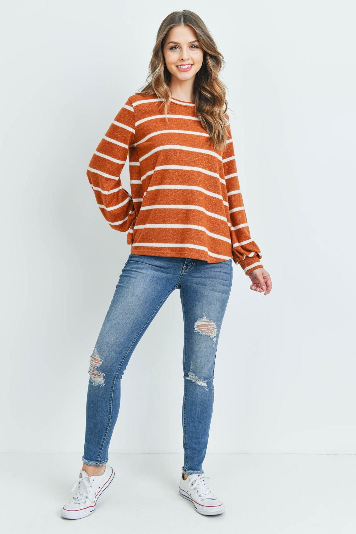 A woman in a Riah Fashion Striped Long Sleeved Round Neck Swing Top and jeans stands against a pink backdrop, wearing a long necklace and touching her hair.