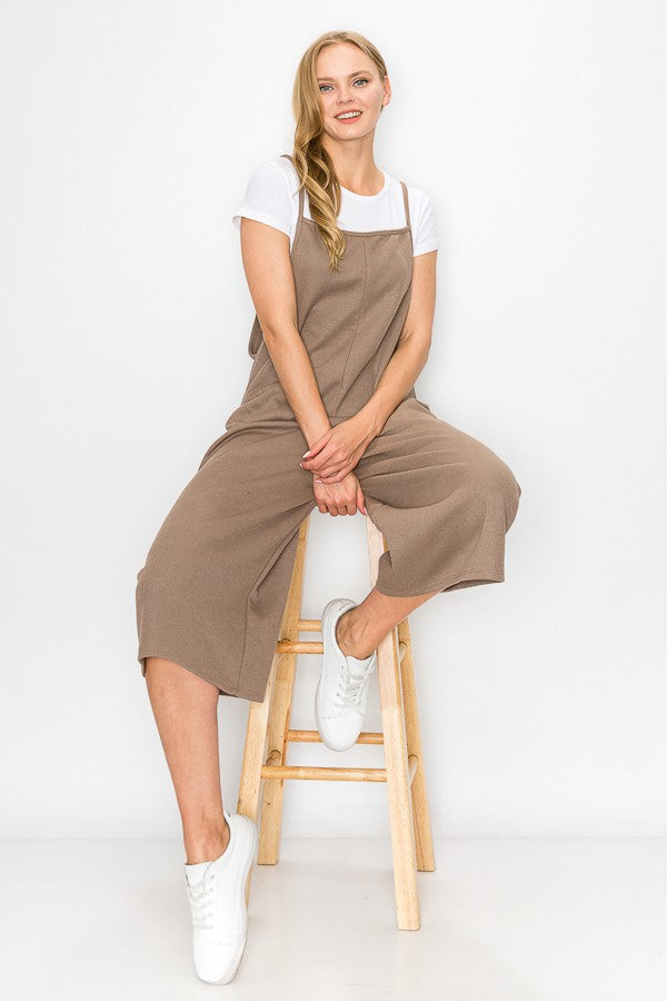 A person with long blonde hair sits on a wooden stool against a plain background, wearing Stylespects Cropped Bottom Wide Leg Oversized Jumpsuit in taupe and white sneakers.