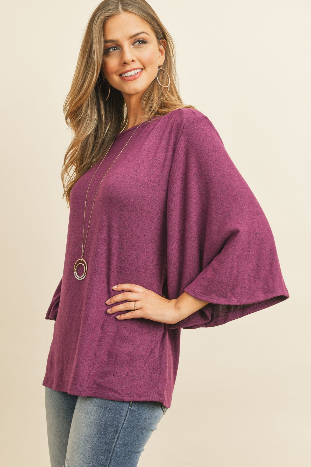 A person in a Riah Fashion Boat Neck Bell Sleeve Solid Hacci Brushed Top and ripped jeans stands against a neutral background, accessorized with hoop earrings and a long necklace.