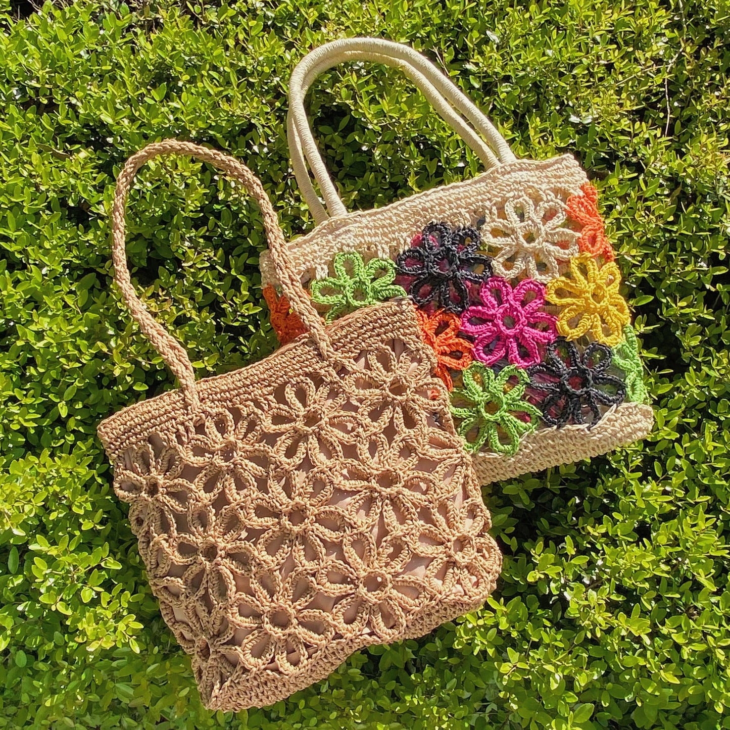 The Hand Made Flower Power Tote by Ellison + Young, featuring colorful floral crochet patterns in orange, green, pink, black, and yellow, rests on dense green foliage.