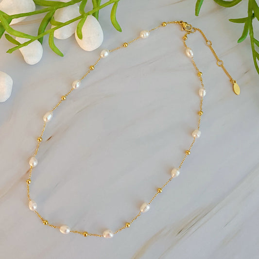 The Ashley Freshwater Pearl Necklace by Ellison + Young is a delicate gold chain adorned with small pearls and tiny gold beads, elegantly displayed on a light marble surface with green leaves and white stones.