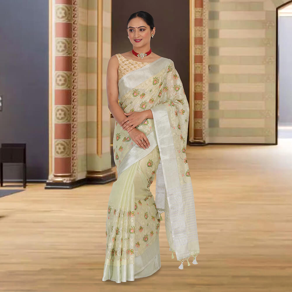 A woman in Chiros Tissue Banarasi Saree With Embroidery - Sea Green stands indoors on a wooden floor, against a decorative wall.