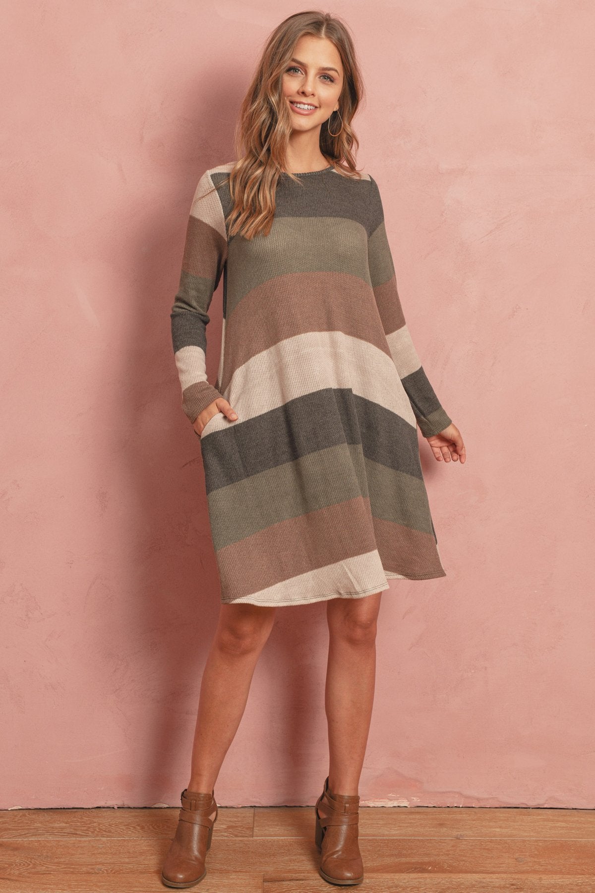 A woman poses in the Riah Fashion Long Sleeved Rib Stripe Pocket Dress in Charcoal/Mocha against a pink textured background.