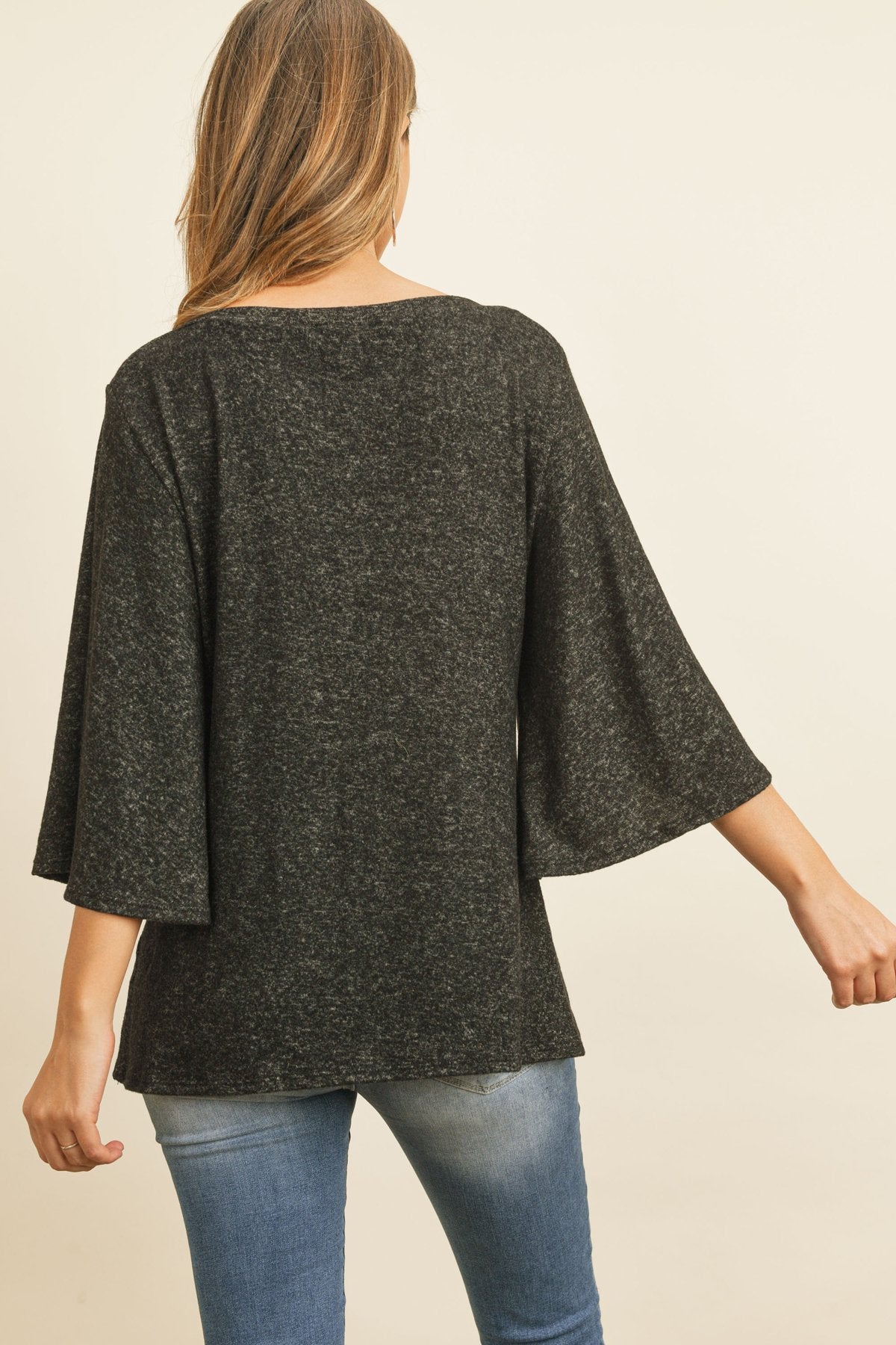 A person in a Riah Fashion Boat Neck Bell Sleeve Solid Hacci Brushed Top and ripped jeans stands against a neutral background, accessorized with hoop earrings and a long necklace.