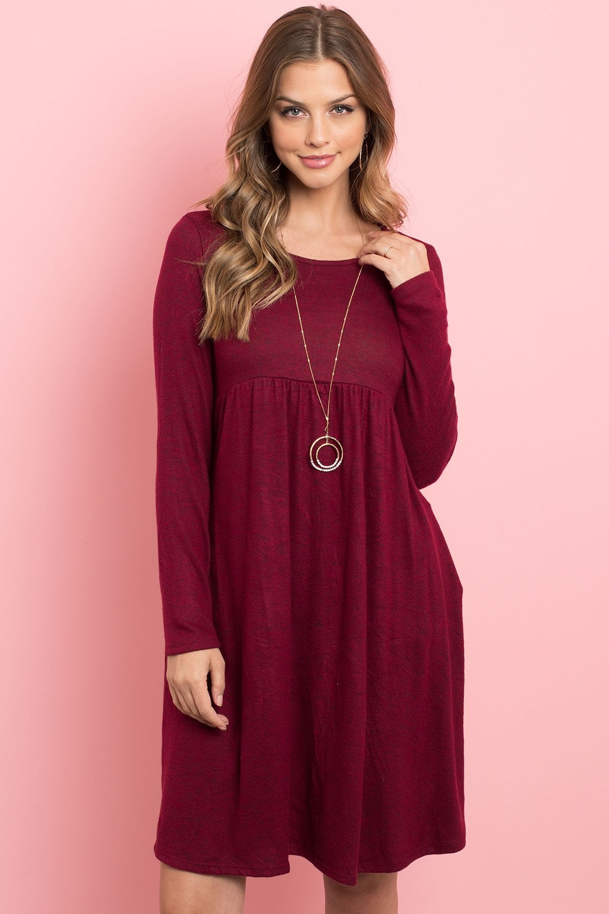 A woman in a Riah Fashion Boat Neck Brushed Hacci Round Hem Dress in Dark Ruby stands before a pink backdrop, adorned with a long necklace featuring circular pendants.