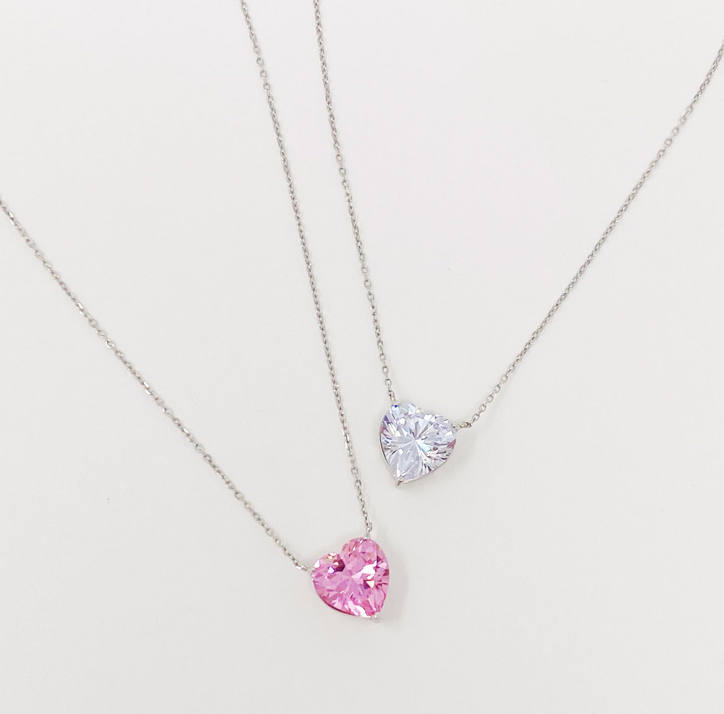 Two Absolute Beauty sterling silver heart necklaces by Ellison + Young; one with a pink gem, the other with a clear gem, against a white background.