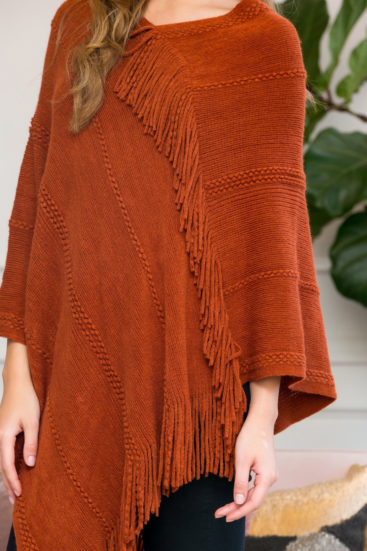 A person stands wearing a Riah Fashion Asymmetric Fringe Poncho in one color, paired with jeans and brown boots, smiling while holding the poncho. In the background are a plant and a sofa with cushions.