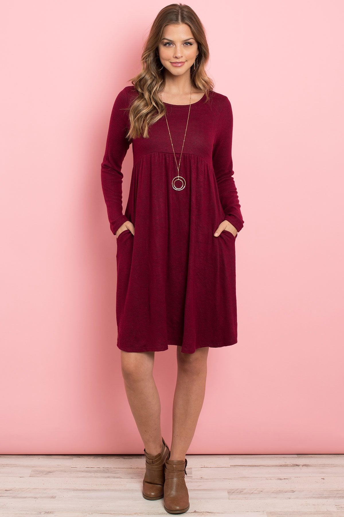 A woman in a Riah Fashion Boat Neck Brushed Hacci Round Hem Dress in Dark Ruby stands before a pink backdrop, adorned with a long necklace featuring circular pendants.