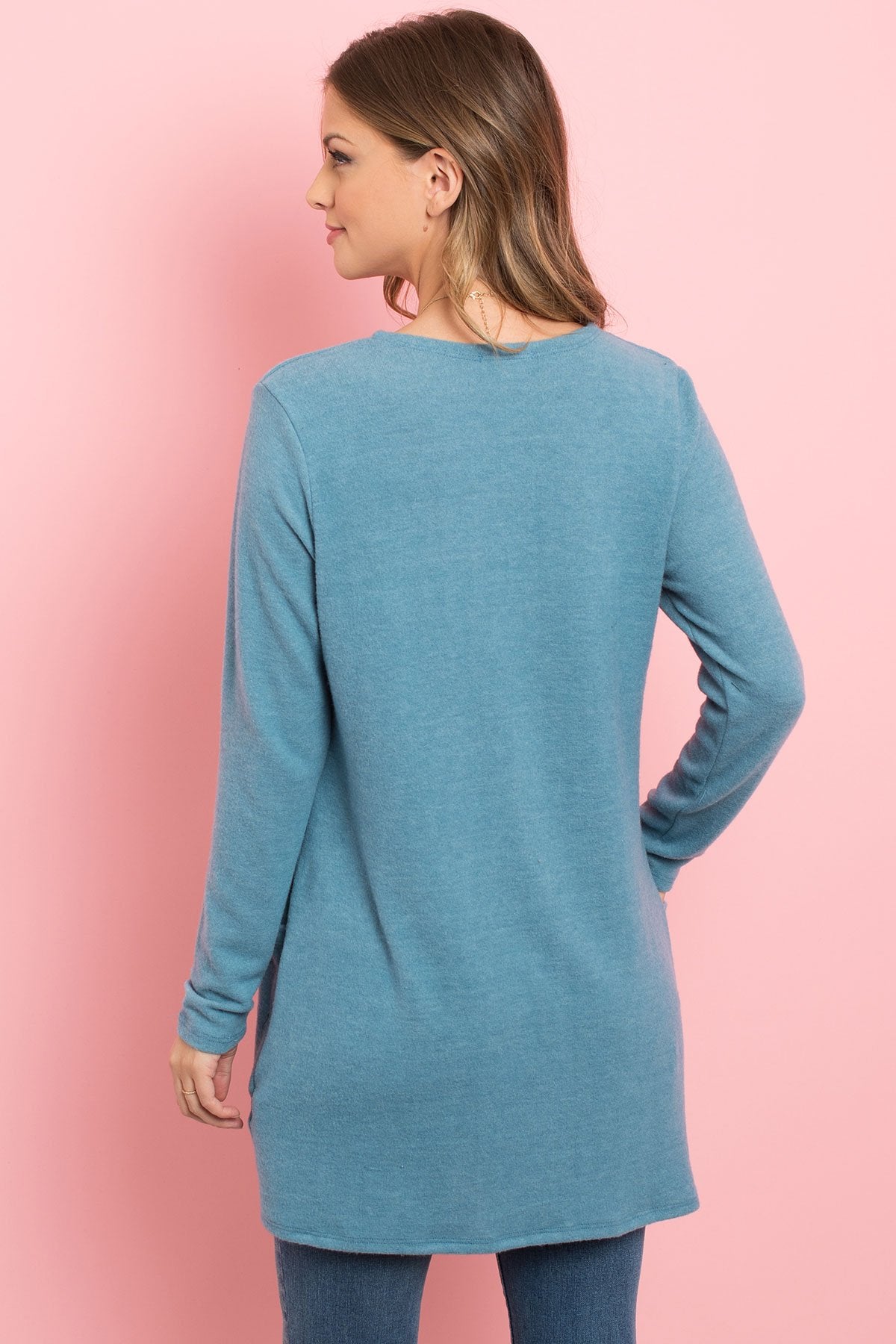 A woman in a teal Solid Long Sleeved Open Front Pocket Cardigan by Riah Fashion over a beige top stands against a pink background, smiling with hands in pockets.