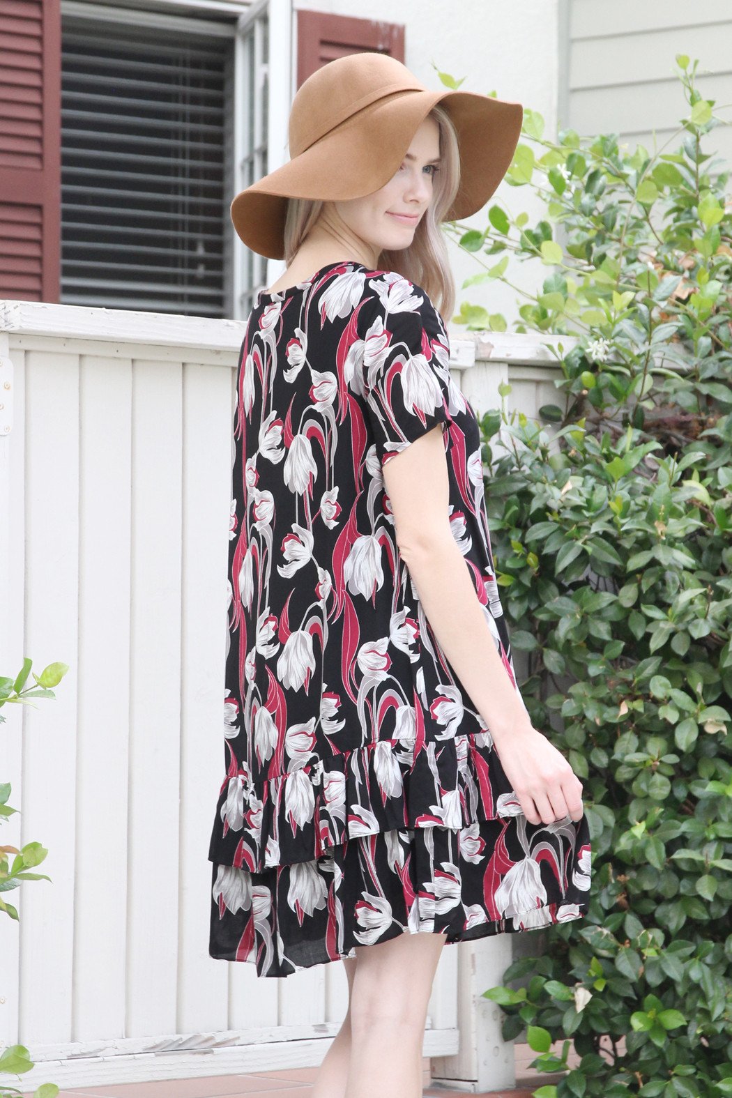 A woman in a Riah Fashion Short Sleeve Floral Ruffle Dress and a brown hat stands near a wooden fence and greenery.