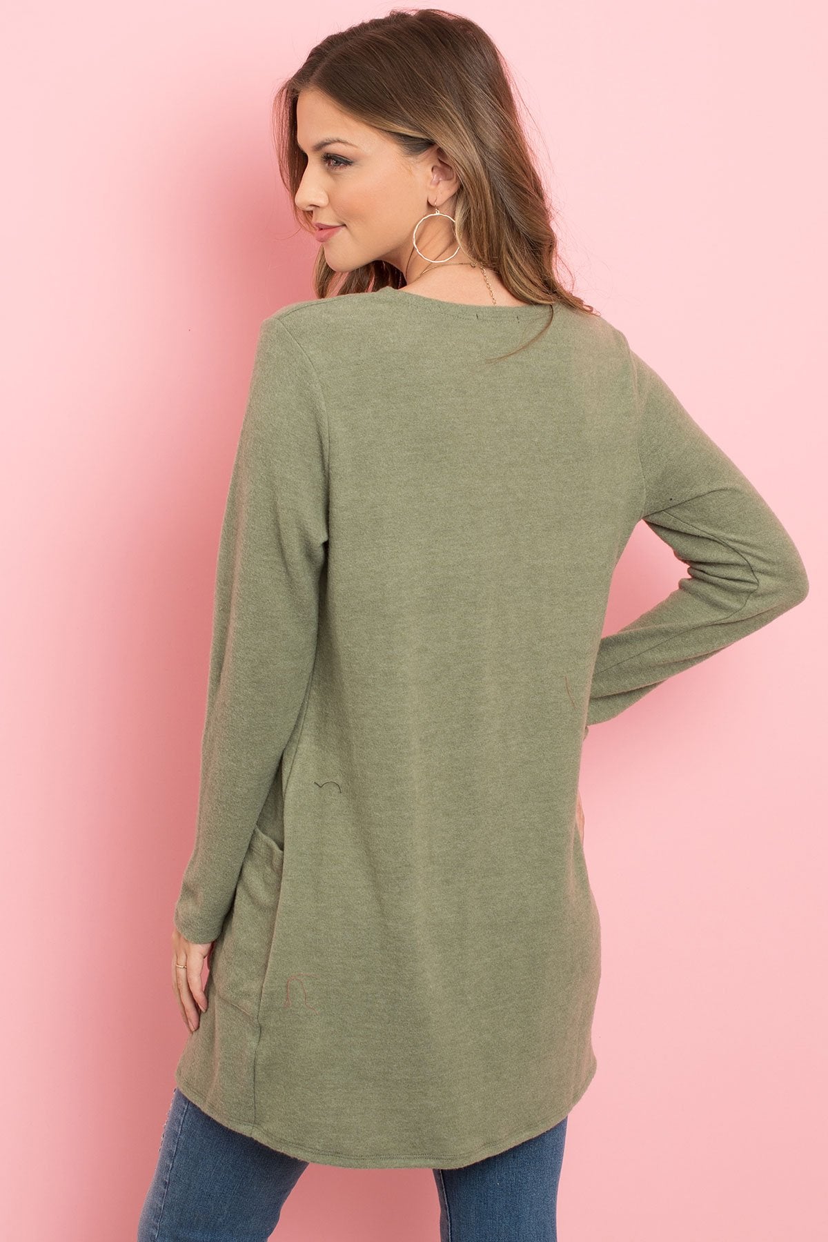 A woman in a teal Solid Long Sleeved Open Front Pocket Cardigan by Riah Fashion over a beige top stands against a pink background, smiling with hands in pockets.