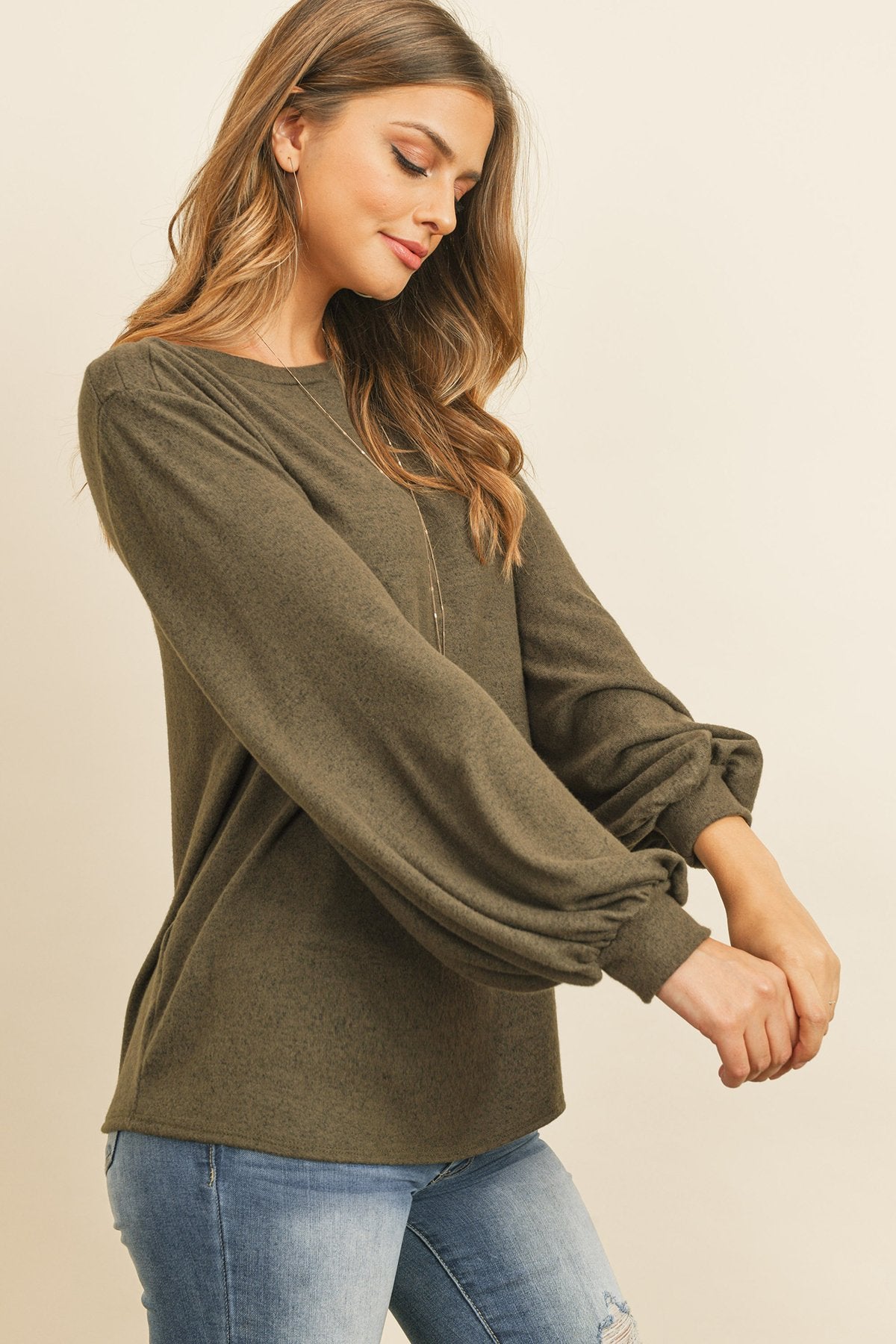 A woman models a Riah Fashion Puff Sleeved Boat Neck Two Toned Brushed Hacci Top with ripped jeans and brown boots on a concrete floor against a plain backdrop, accessorized with a long necklace.