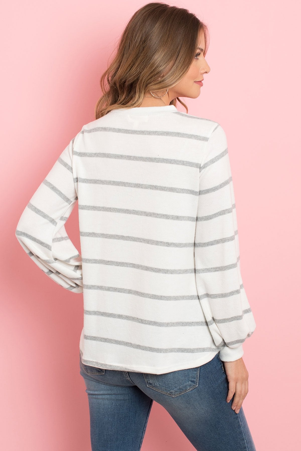 A woman in a Riah Fashion Striped Long Sleeved Round Neck Swing Top and jeans stands against a pink backdrop, wearing a long necklace and touching her hair.