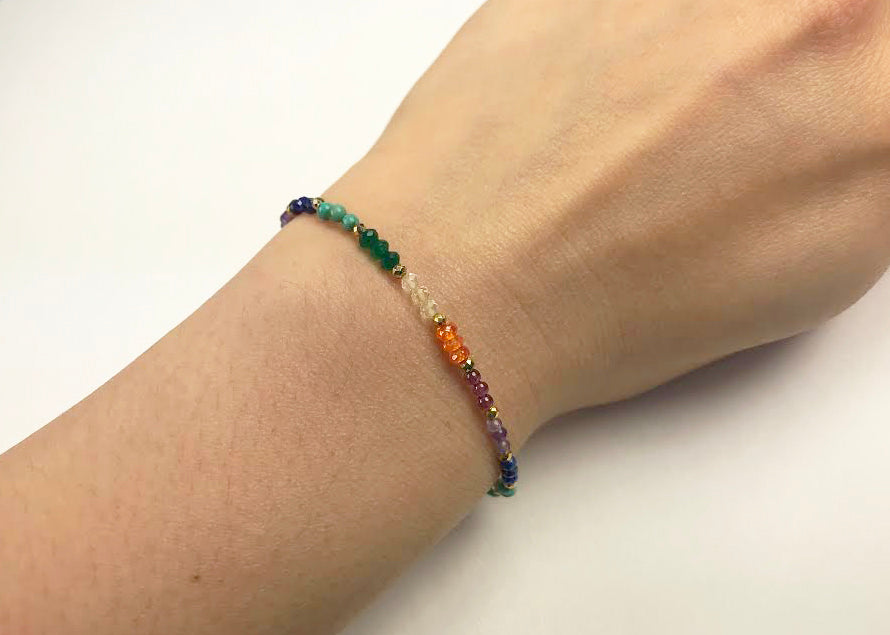 The Chakra Delicate Bracelet by Gena Myint is a colorful, beaded bracelet with a gold chain and a heart-shaped charm, featuring small beads in orange, purple, green, and blue.
