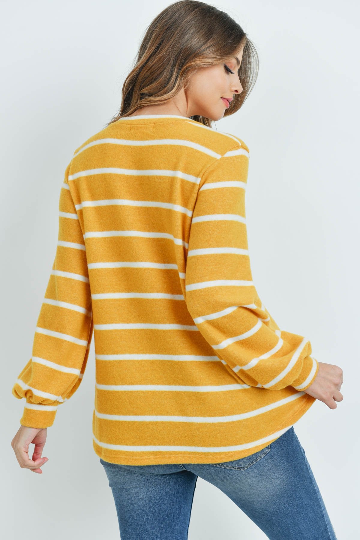 A woman in a Riah Fashion Striped Long Sleeved Round Neck Swing Top and jeans stands against a pink backdrop, wearing a long necklace and touching her hair.