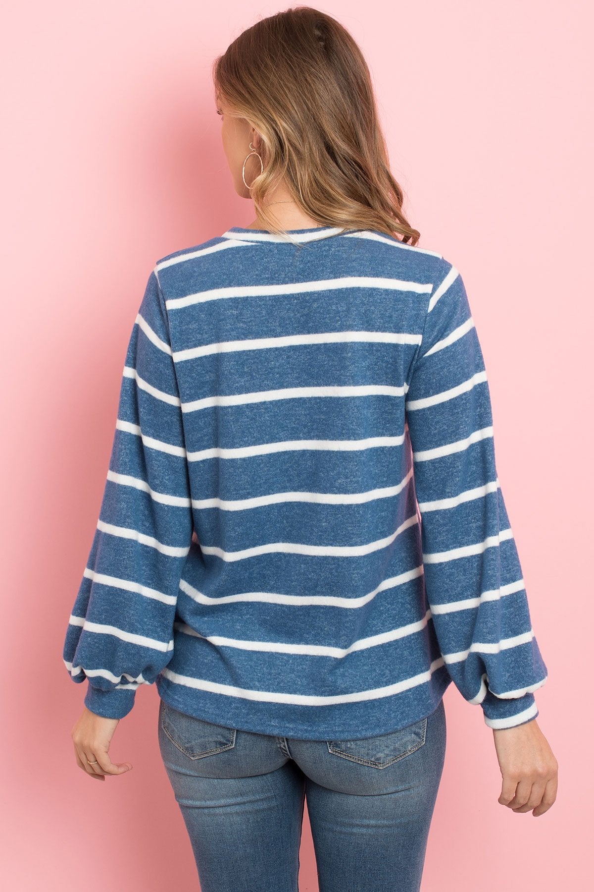 A woman in a Riah Fashion Striped Long Sleeved Round Neck Swing Top and jeans stands against a pink backdrop, wearing a long necklace and touching her hair.