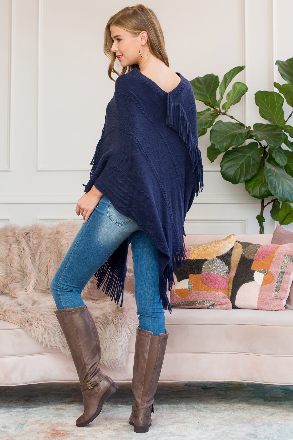 A person stands wearing a Riah Fashion Asymmetric Fringe Poncho in one color, paired with jeans and brown boots, smiling while holding the poncho. In the background are a plant and a sofa with cushions.