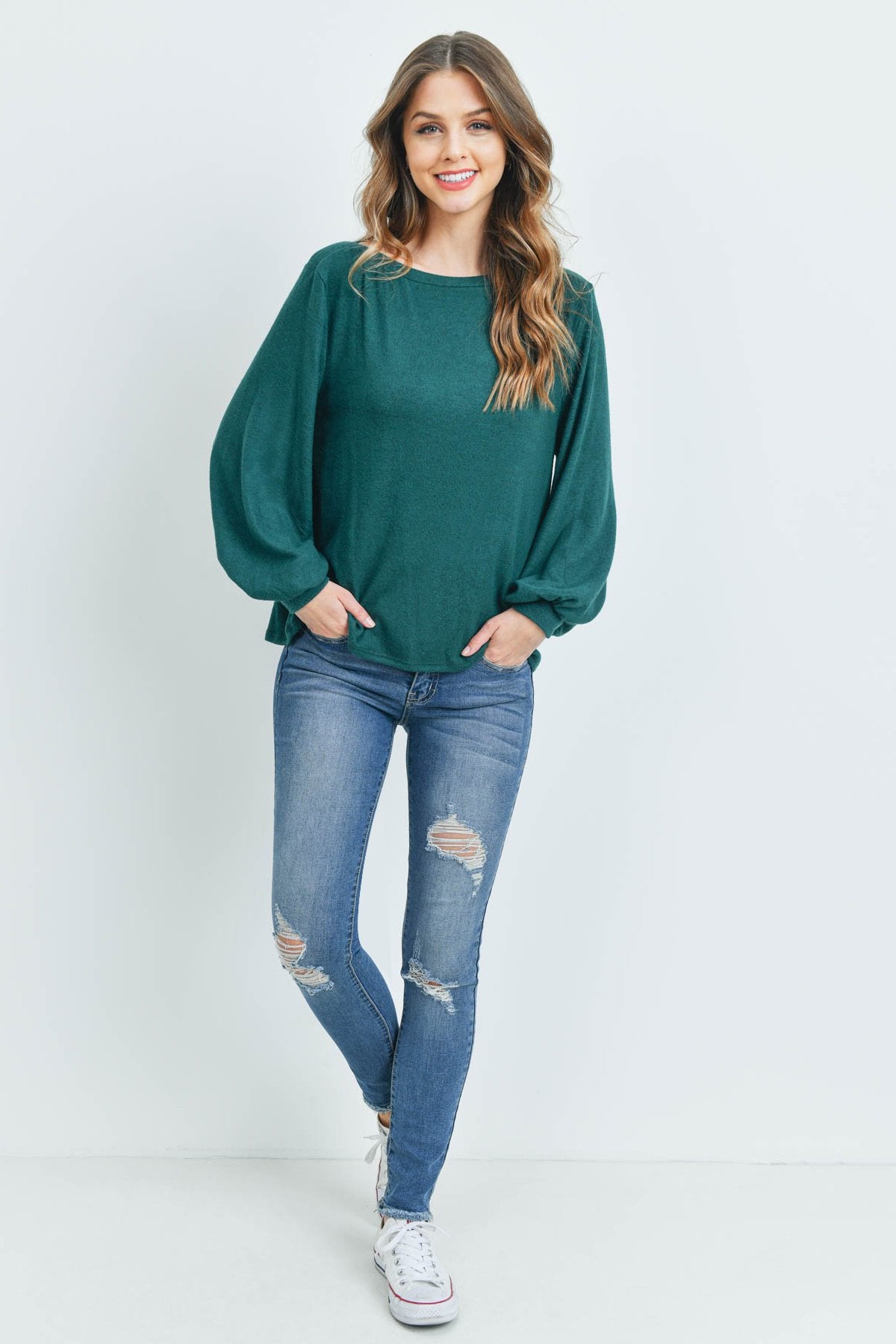 A woman models a Riah Fashion Puff Sleeved Boat Neck Two Toned Brushed Hacci Top with ripped jeans and brown boots on a concrete floor against a plain backdrop, accessorized with a long necklace.