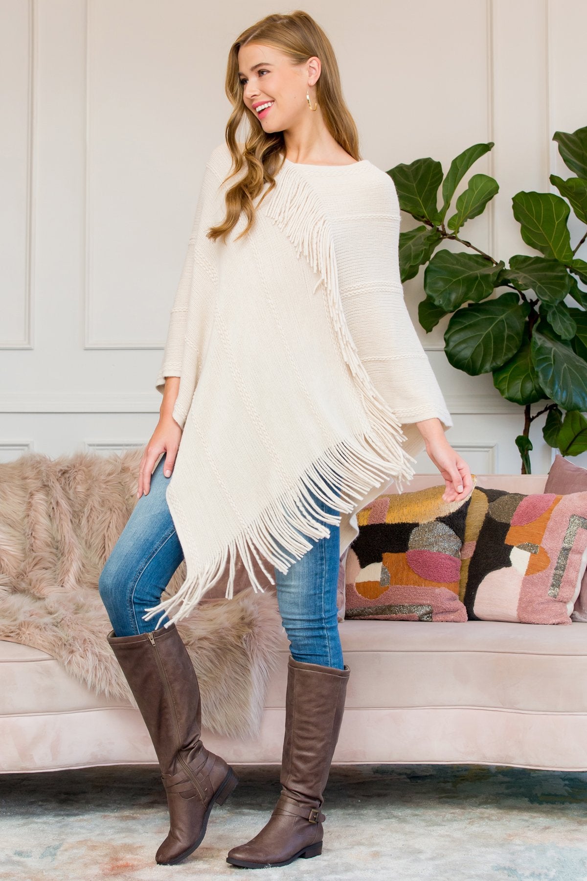 A person stands wearing a Riah Fashion Asymmetric Fringe Poncho in one color, paired with jeans and brown boots, smiling while holding the poncho. In the background are a plant and a sofa with cushions.