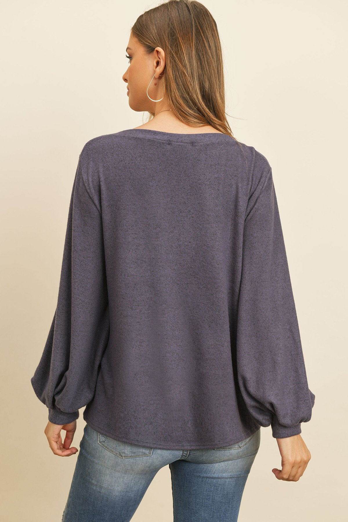 A woman models a Riah Fashion Puff Sleeved Boat Neck Two Toned Brushed Hacci Top with ripped jeans and brown boots on a concrete floor against a plain backdrop, accessorized with a long necklace.