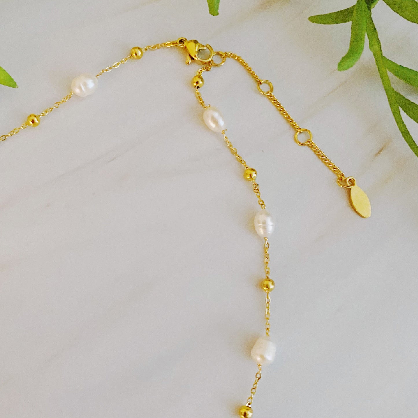 The Ashley Freshwater Pearl Necklace by Ellison + Young is a delicate gold chain adorned with small pearls and tiny gold beads, elegantly displayed on a light marble surface with green leaves and white stones.