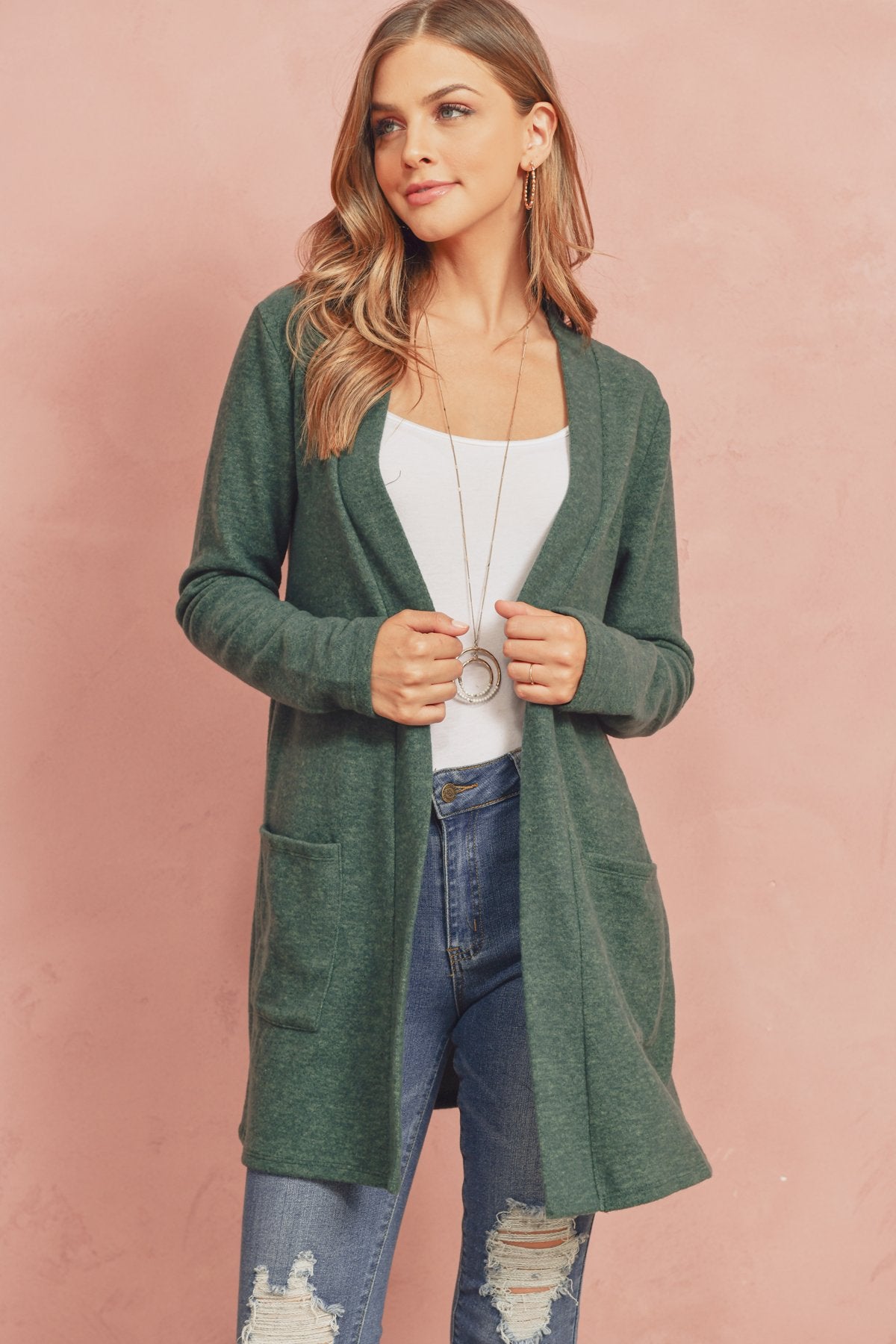 A woman in a Brushed Melange Open Front Cardigan by Riah Fashion and ripped jeans stands against a pink background, hands in her pockets.