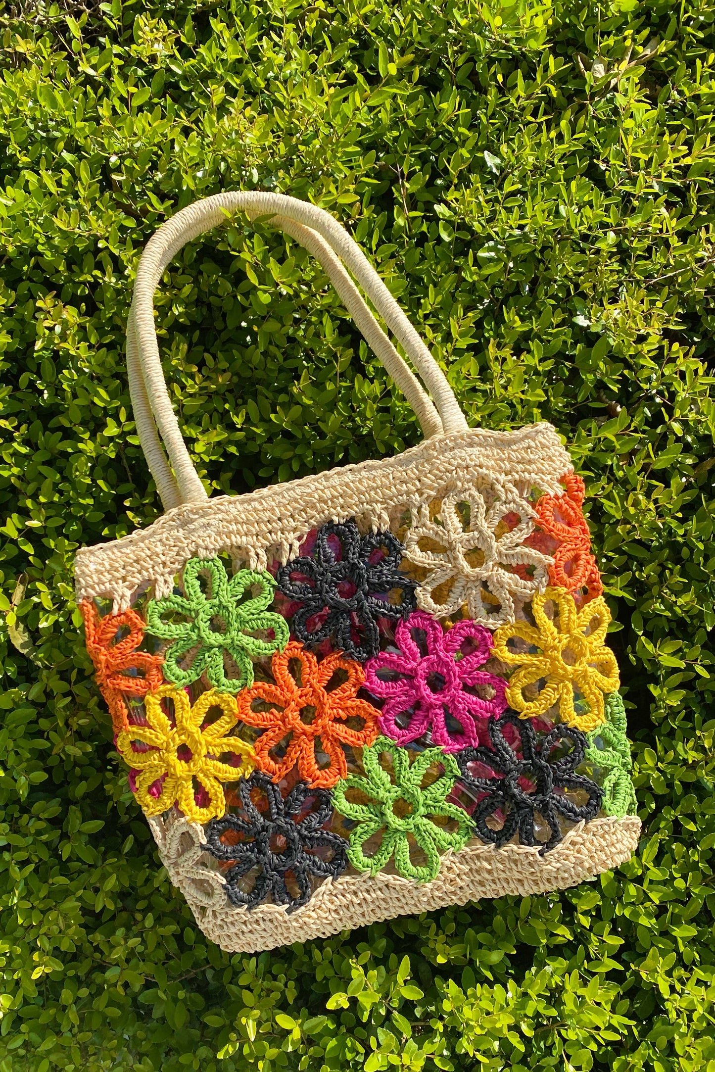 The Hand Made Flower Power Tote by Ellison + Young, featuring colorful floral crochet patterns in orange, green, pink, black, and yellow, rests on dense green foliage.