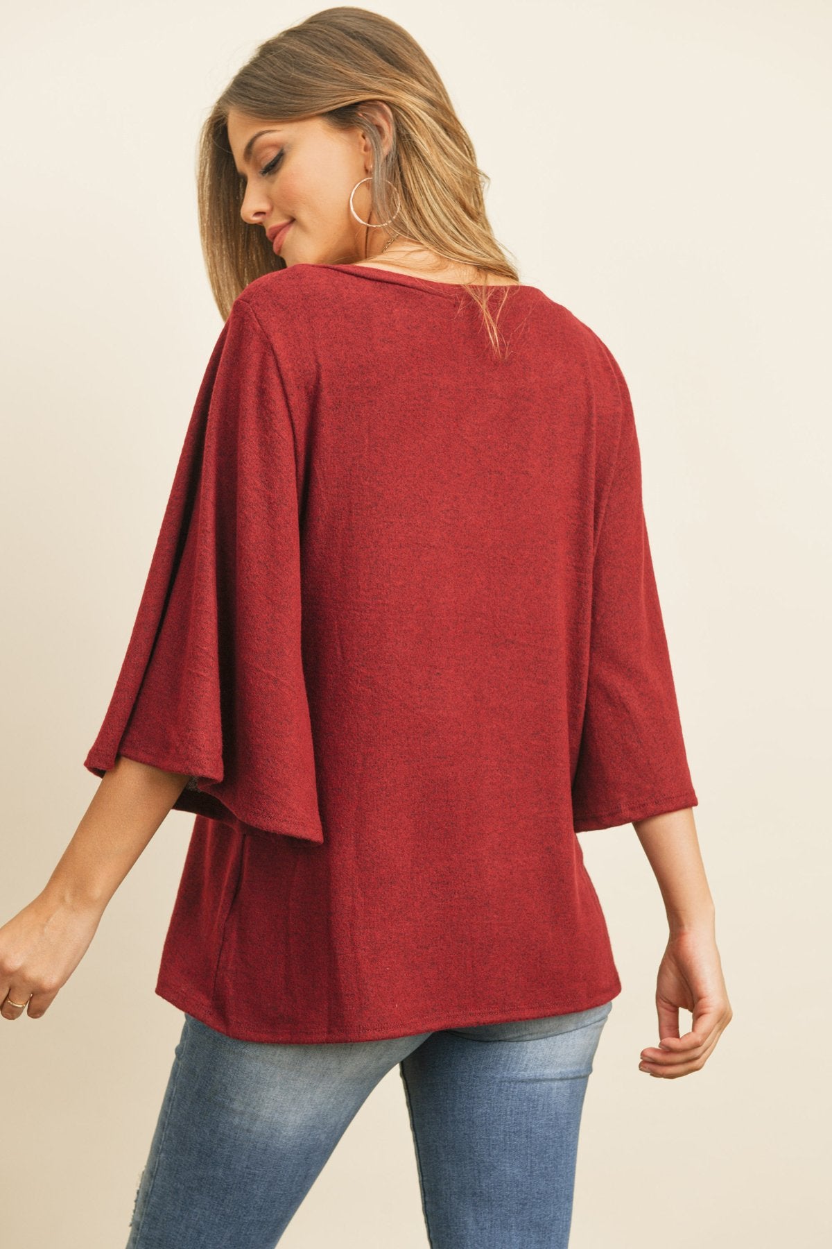 A person in a Riah Fashion Boat Neck Bell Sleeve Solid Hacci Brushed Top and ripped jeans stands against a neutral background, accessorized with hoop earrings and a long necklace.
