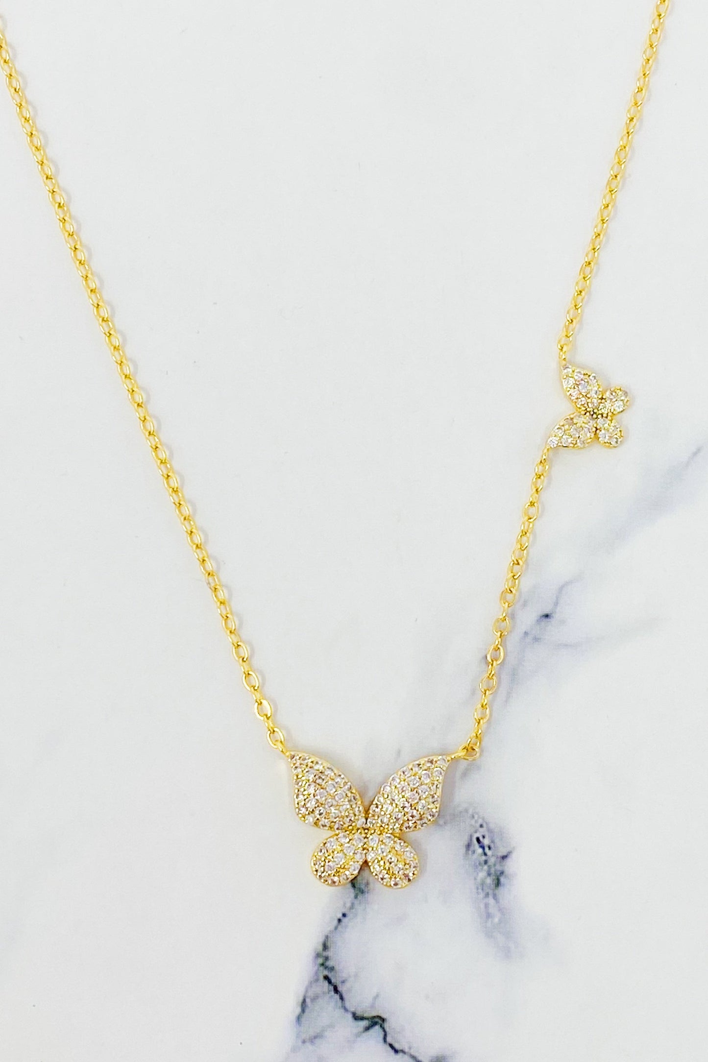 The Ellison + Young Double Shine Butterfly Necklace, featuring butterfly-shaped pendants adorned with small crystals, is elegantly displayed on a light marble background.
