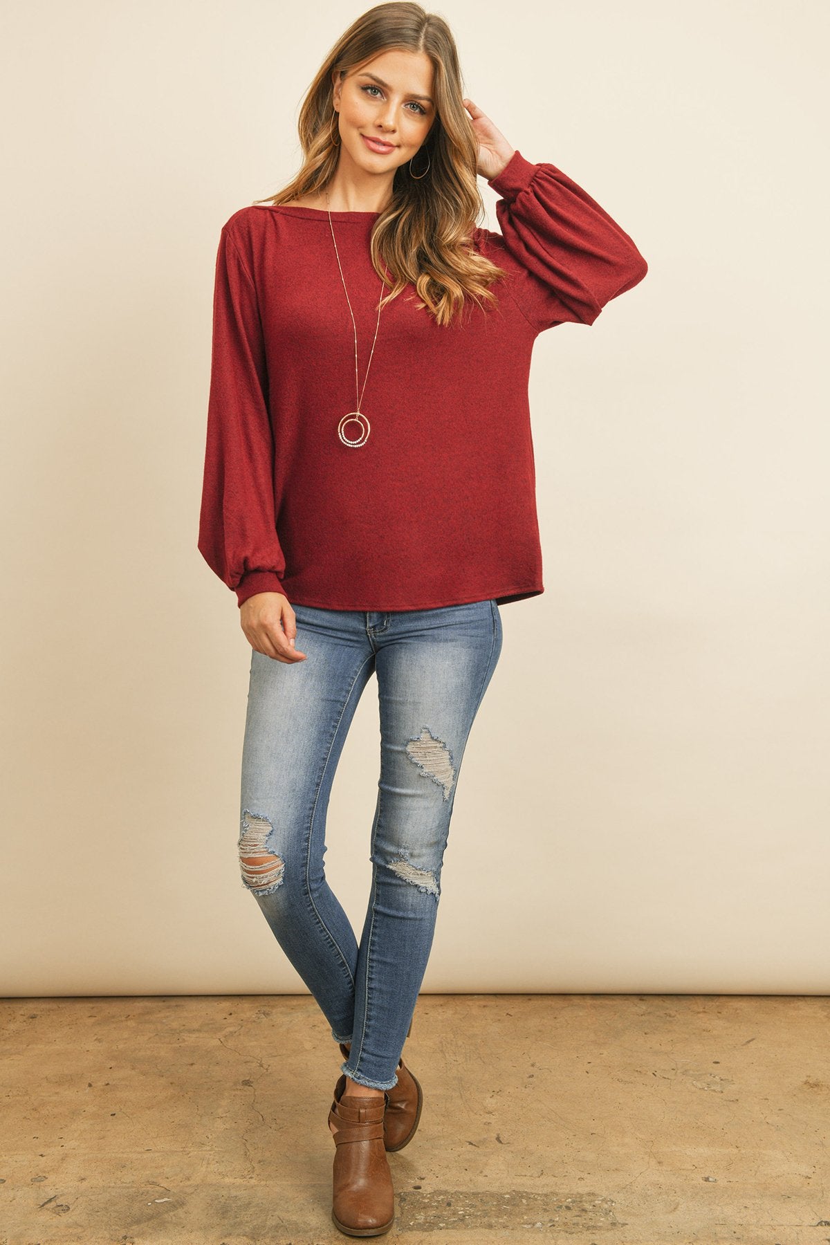 A woman models a Riah Fashion Puff Sleeved Boat Neck Two Toned Brushed Hacci Top with ripped jeans and brown boots on a concrete floor against a plain backdrop, accessorized with a long necklace.