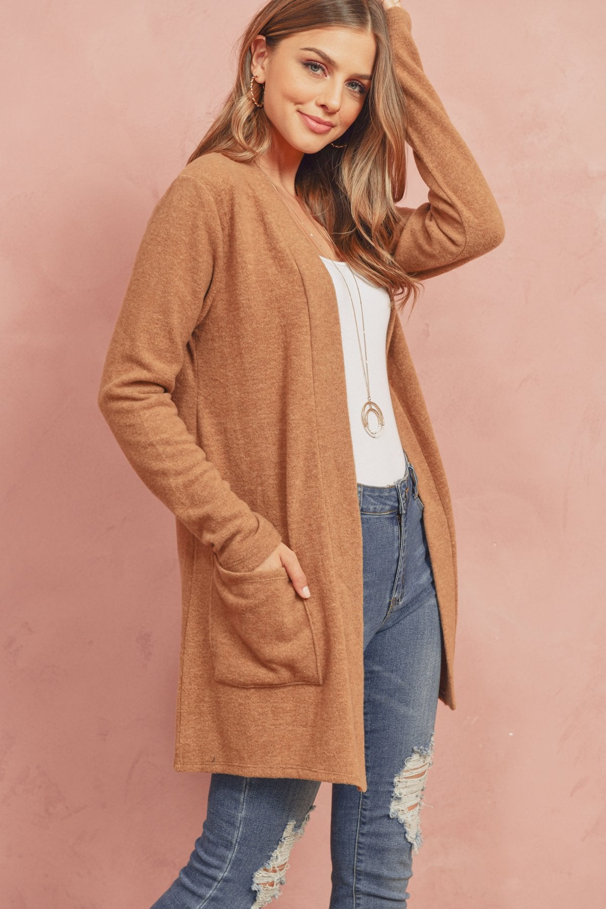 A woman in a Brushed Melange Open Front Cardigan by Riah Fashion and ripped jeans stands against a pink background, hands in her pockets.