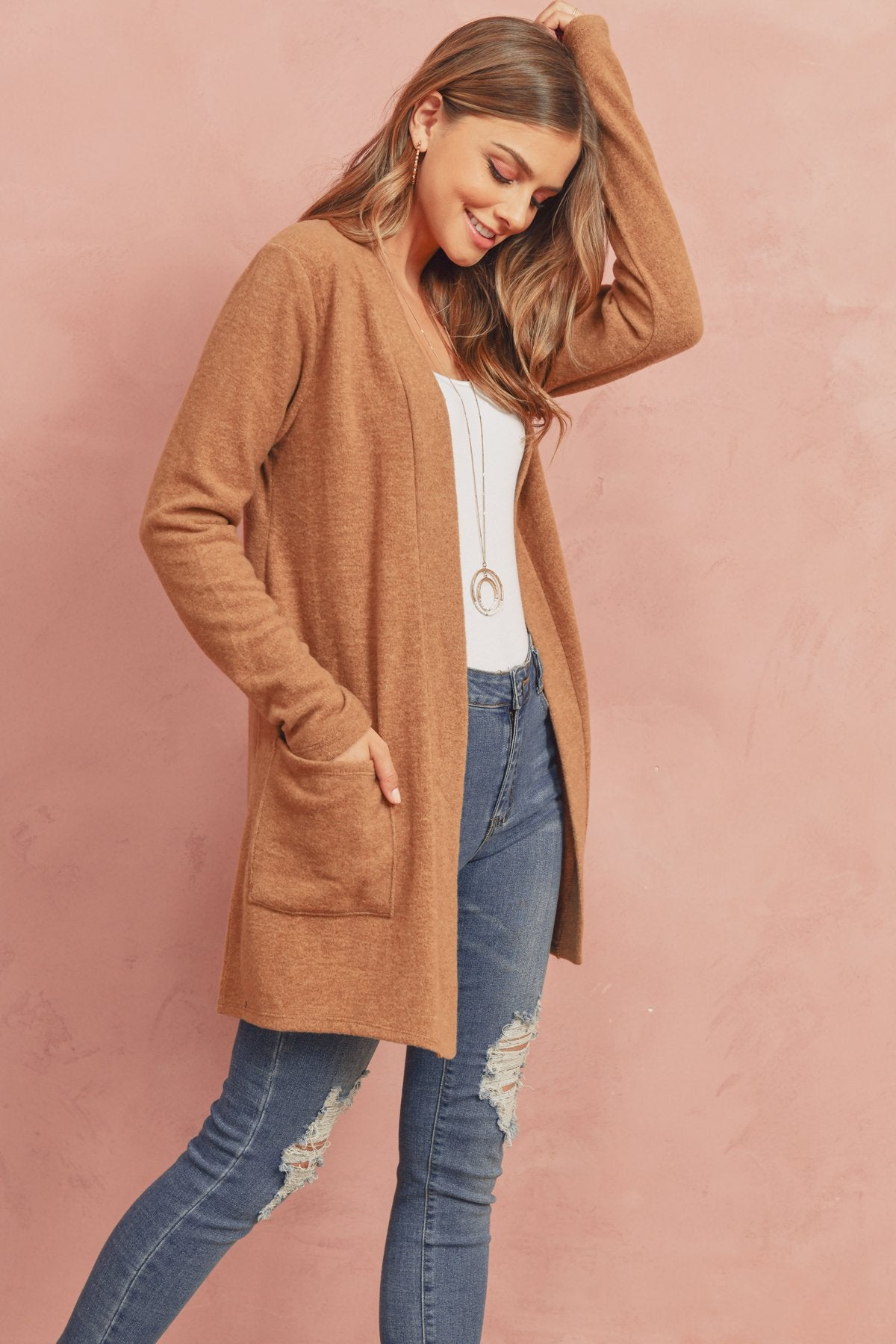 A woman in a Brushed Melange Open Front Cardigan by Riah Fashion and ripped jeans stands against a pink background, hands in her pockets.