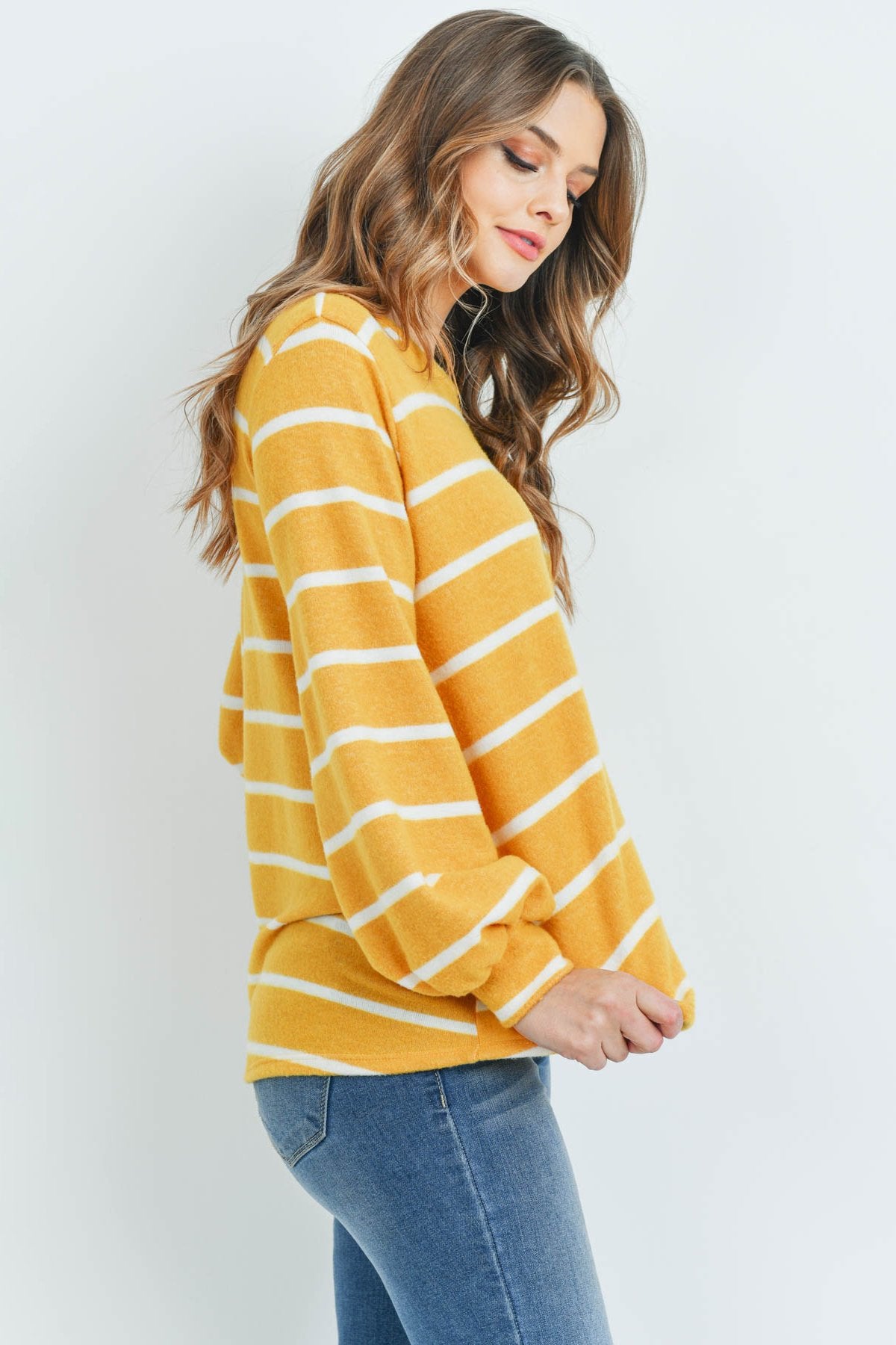 A woman in a Riah Fashion Striped Long Sleeved Round Neck Swing Top and jeans stands against a pink backdrop, wearing a long necklace and touching her hair.