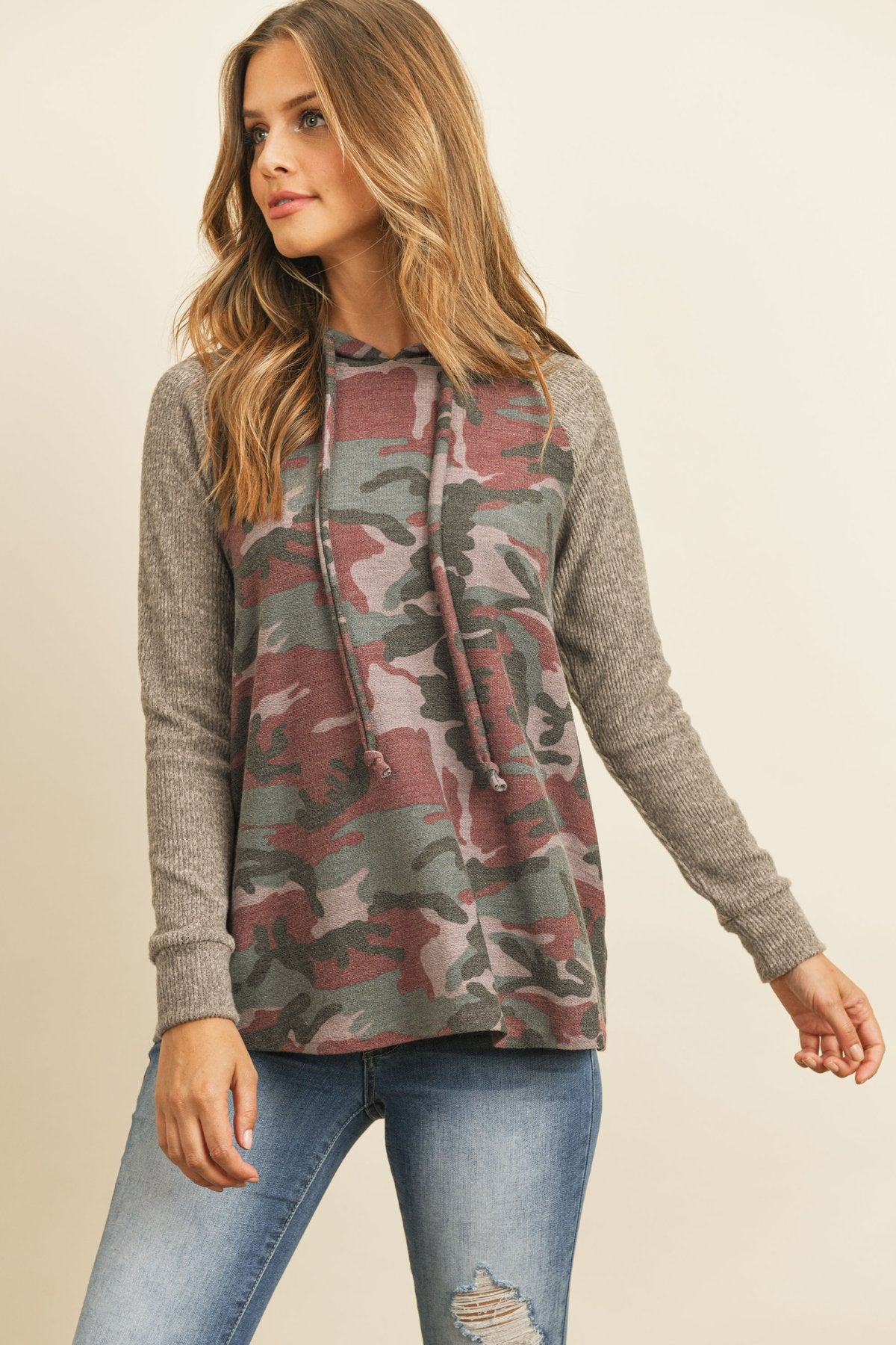 A person wearing the Self Tie Rib Hacci Brushed Sleeve Camouflage Hoodie by Riah Fashion with distressed jeans, holding the hoodie strings, stands against a beige background.