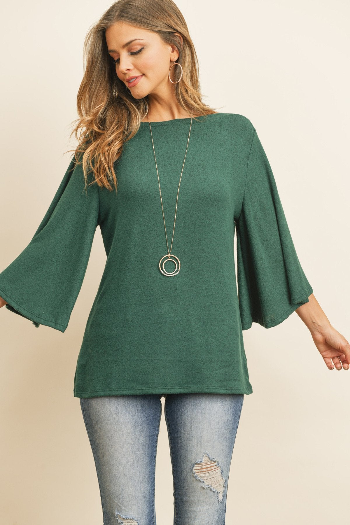 A person in a Riah Fashion Boat Neck Bell Sleeve Solid Hacci Brushed Top and ripped jeans stands against a neutral background, accessorized with hoop earrings and a long necklace.