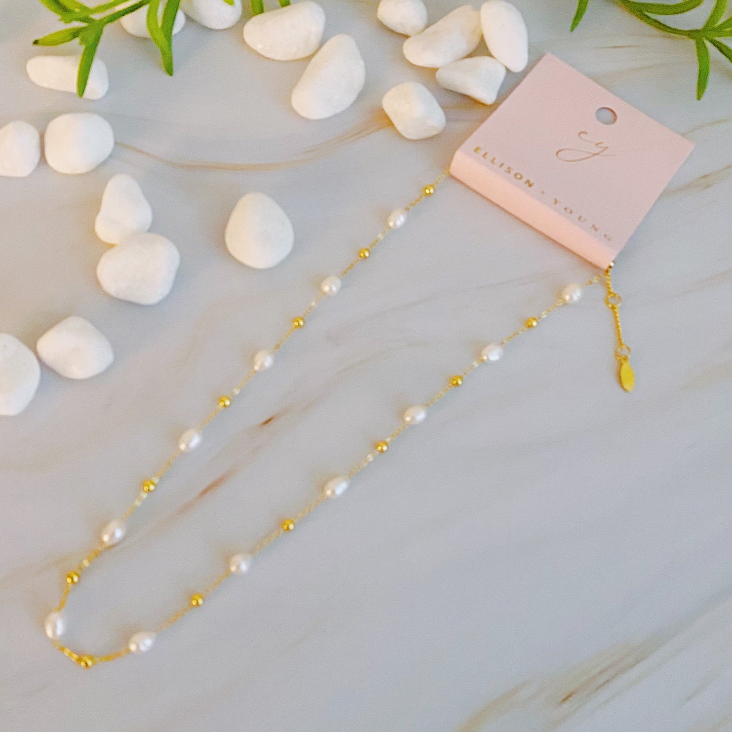 The Ashley Freshwater Pearl Necklace by Ellison + Young is a delicate gold chain adorned with small pearls and tiny gold beads, elegantly displayed on a light marble surface with green leaves and white stones.
