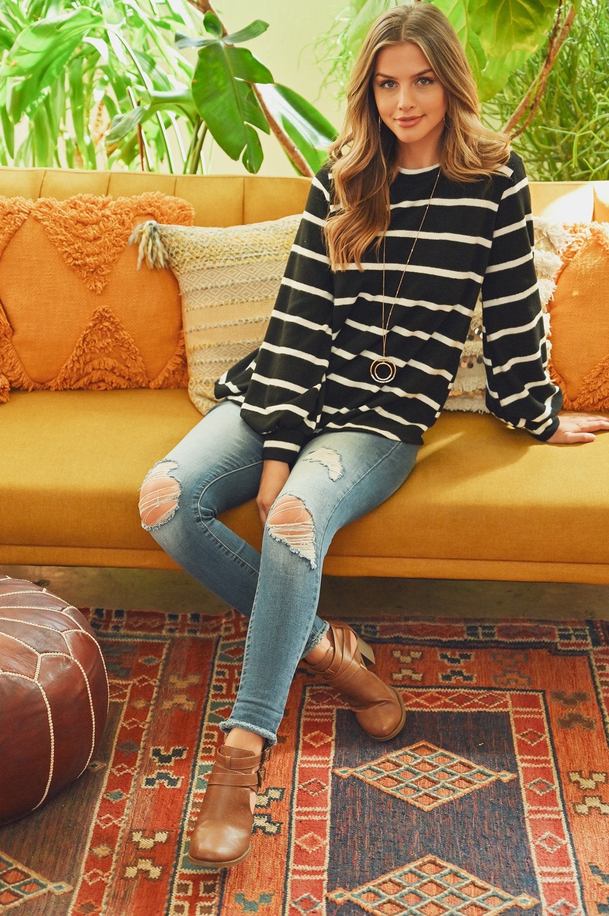 A woman in a Riah Fashion Striped Long Sleeved Round Neck Swing Top and jeans stands against a pink backdrop, wearing a long necklace and touching her hair.